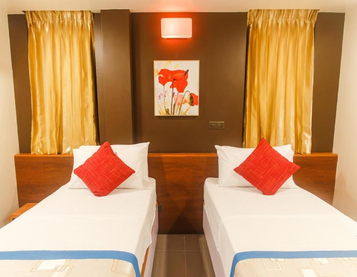 A hotel room with two single beds, each with a white bedspread and a red pillow. There is a wall-mounted light above the beds and a painting of red flowers on the wall.