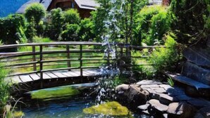 A rustic wooden bridge over a clear stream with a waterfall, surrounded by lush greenery and houses in the background. - st martin chalets by Interhome (Sankt Michael im Lungau)