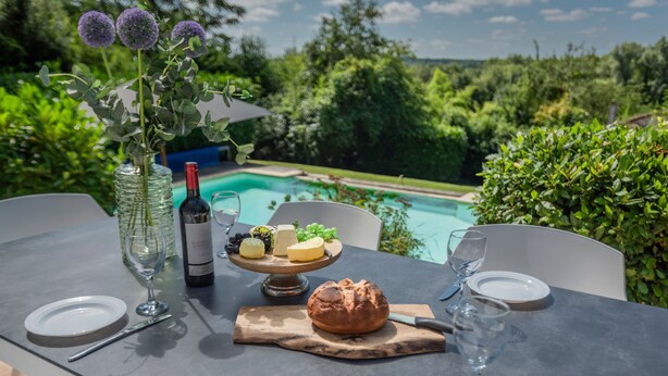 Ein Essplatz im Freien mit einer Weinflasche, Käse, Trauben und Brot, vor einer Kulisse aus Schwimmbecken und Grünflächen.