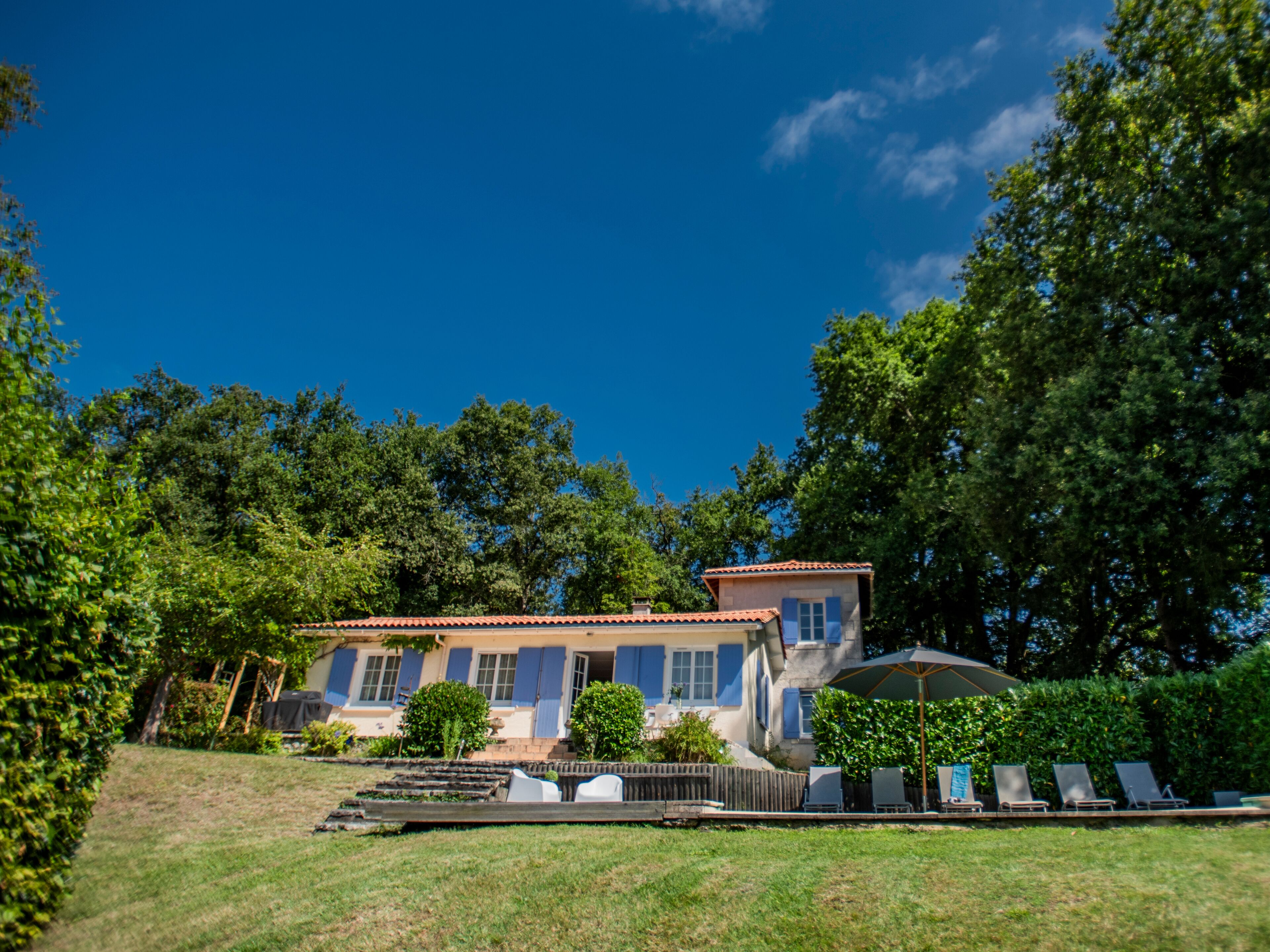 Une maison à deux étages, avec un toit recouvert de tuiles rouges, un porche à l’avant doté d’une balustrade en bois, et un petit jardin agrémenté d’une terrasse en bois et de mobilier d’extérieur.