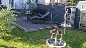 Un patio trasero con una terraza de madera, una mesa con sillas, un columpio colgante y un pequeño jardín.