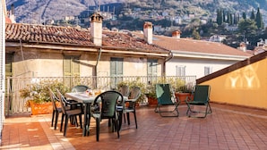 Terraço com mesa e cadeiras, com vista para uma vila no topo de uma colina, com edifícios e árvores.