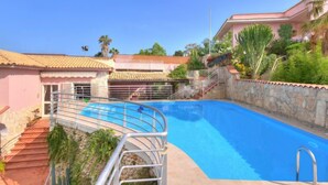 Une piscine entourée d’une rambarde métallique, elle-même bordée d’un mur de pierre et d’un escalier menant à un bâtiment dont le toit est recouvert de tuiles.