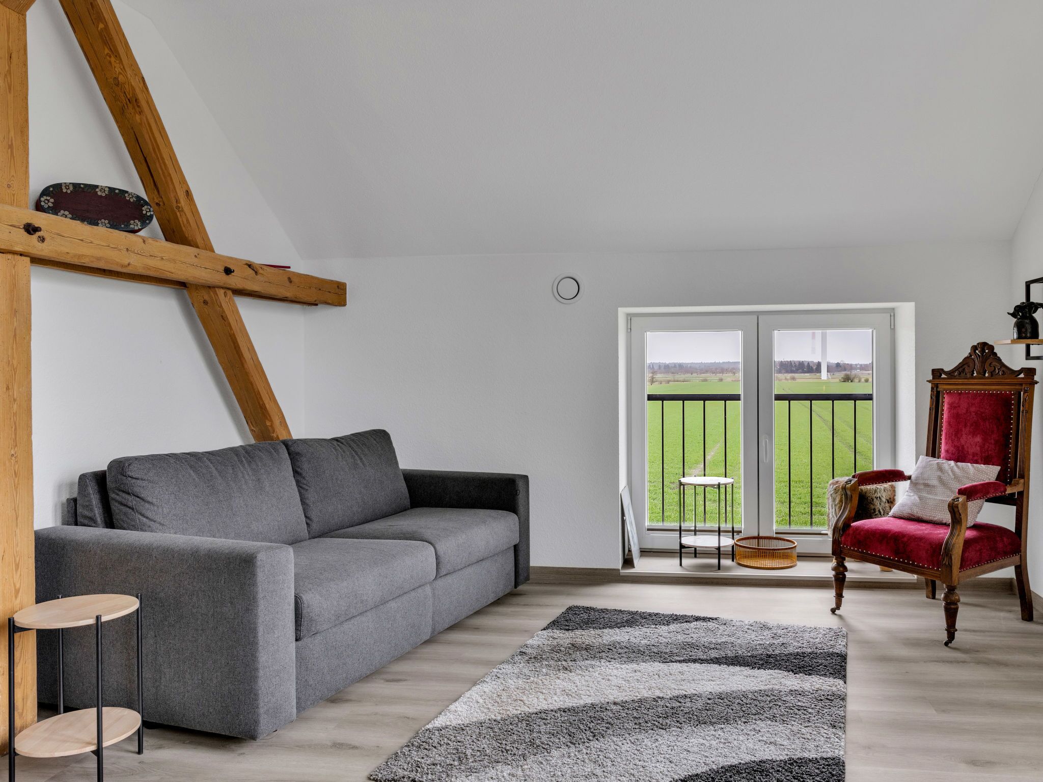 A modern living room with a grey sofa, a red chair, a patterned rug, and a balcony with a view of a green field.