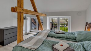 Ein Schlafzimmer mit schrägverdeckter Decke, Holzbalken und einem Bett mit grüner Bettwäsche, mit Blick auf einen Garten durch eine Glastür.