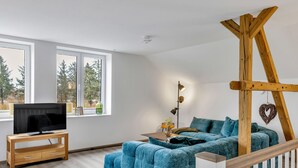 Ein modernes Wohnzimmer mit einer Eckcouch, einem Fernseher auf einem Holzbodenständer, einer Stehlampe und einem Holztrellis mit einem hängenden Herz-Ornament.