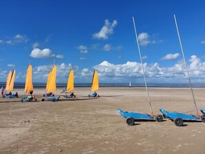 Una playa con varios pequeños veleros de velas naranjas y cascos azules alineados en la arena.