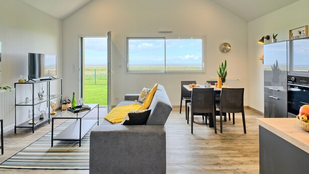 A spacious living area with a grey sofa, a dining table with chairs, a kitchen area, and a large window offering a view of the outdoors. - l'atelier des rêves by Interhome (Cherrueix)