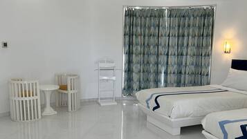 A modern hotel room with a large bed, a small table, two cribs, and a window with curtains.