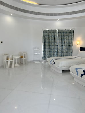 A modern hotel room with a large bed, a small table, two cribs, and a window with curtains. - Ashok Vatika Ananta Elite  Ranakpur (Desuri)