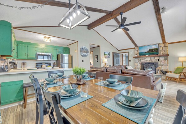 A spacious living area with a wooden dining table set for four, a green kitchen with modern appliances, a fireplace, and a ceiling fan.