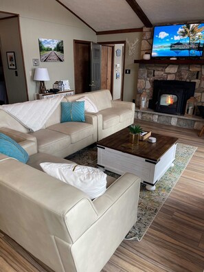 A cozy living room with a beige sofa, a coffee table, a fireplace, and a television mounted on the wall. - Lakefront, Hot Tub, Dog Friendly, Resort Amenities (Gouldsboro)