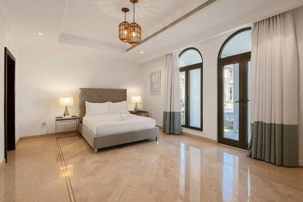 A spacious hotel room with a large bed, a nightstand, a lamp, and a view through arched windows.