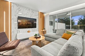 A modern living room with a large flat-screen TV, a grey sofa, a brown armchair, and a round wooden coffee table.