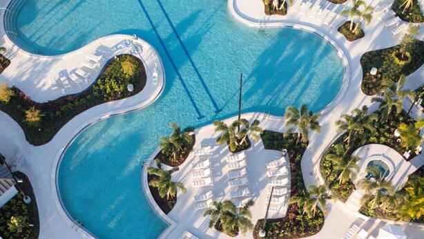 An aerial view of a resort with a large central pool, multiple smaller pools, and surrounding lounge areas with palm trees.