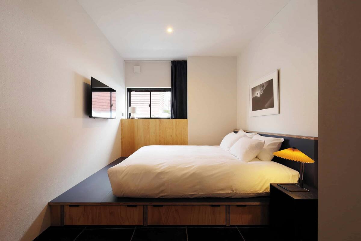 Un dormitorio con cama, mesita de noche, lámpara, ventana y una obra de arte en la pared.