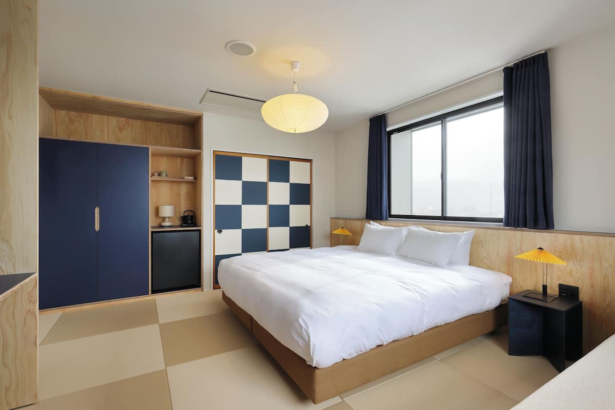 Una habitación de hotel moderna con una cama grande, un armario, un escritorio pequeño y una ventana con cortinas.