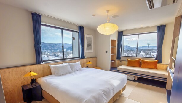 Una habitación de hotel moderna con una cama grande, una mesita de noche con lámpara, un banco con cojines y ventanas amplias con vistas a la ciudad.