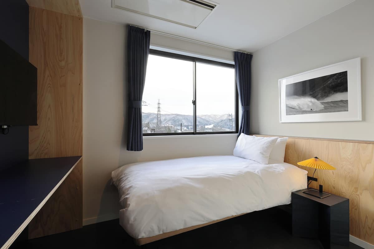 Una habitación de hotel con una cama, una mesita de noche con lámpara, una ventana con cortinas y un cuadro enmarcado en la pared.