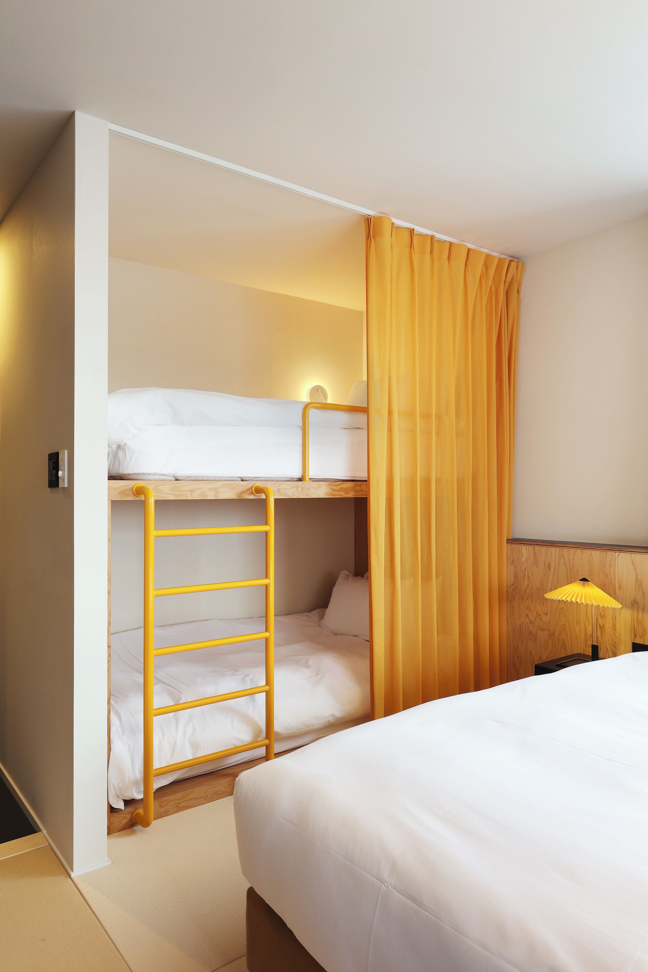 A hotel room with a bunk bed, a ladder, a bed, a wooden headboard, and a yellow lamp.
