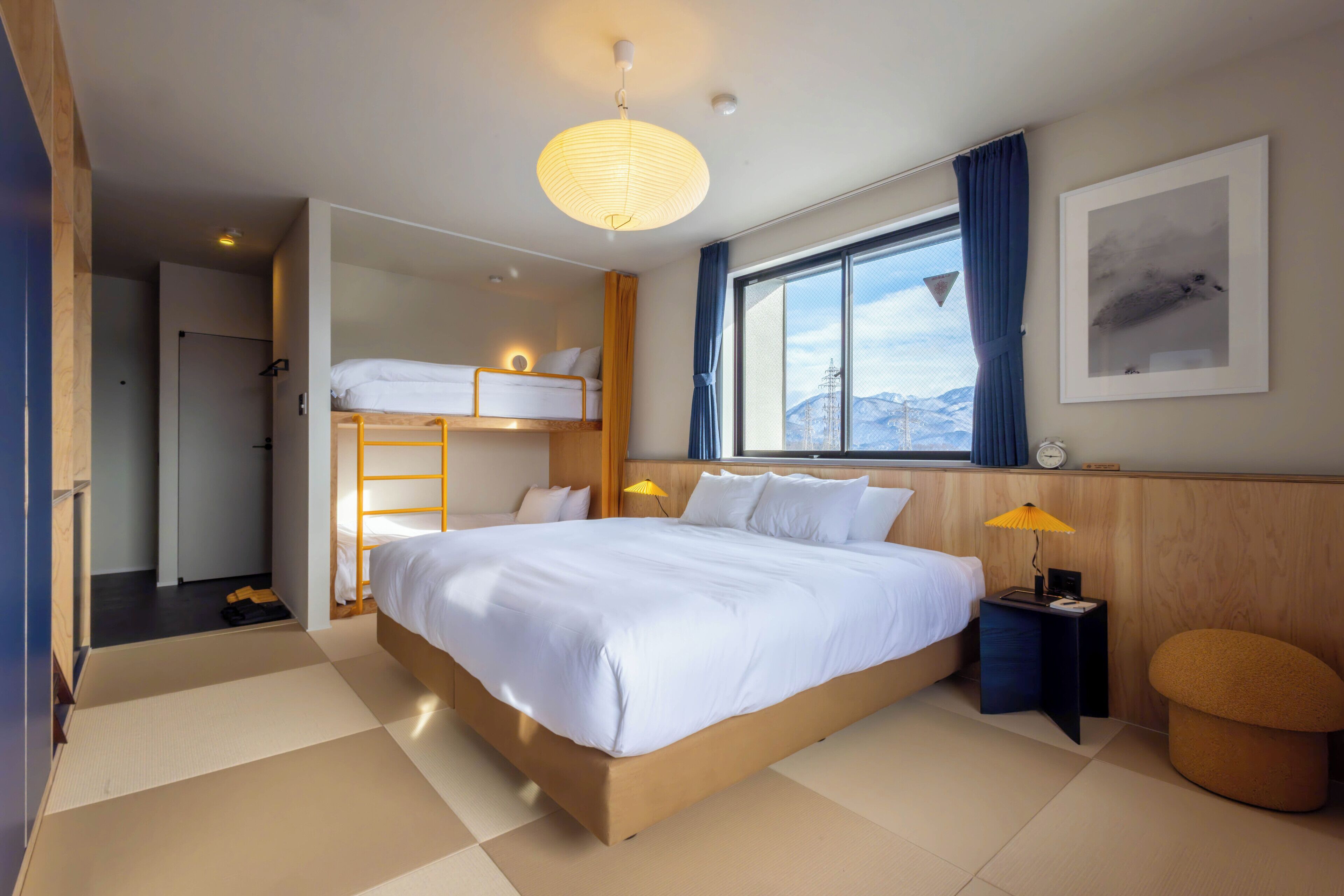 A modern hotel room with a large bed, a window with a view of mountains, a bedside table with a lamp, and a small stool.