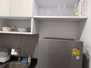 Una cocina compacta con un refrigerador de acero inoxidable, un lavamanos con grifo y un estante superior con algunos objetos.