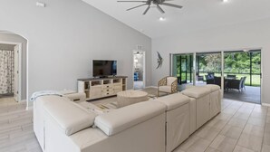Ein großzügiges Wohnzimmer mit einem großen, weißen Modulsofa, einem Flachbildfernseher und einem Deckenventilator. Eine Glastür führt zu einer Außenpatiofläche.