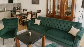Een woonkamer met een groene bank, een bijpassende fauteuil en poef, een houten salontafel, en een eethoek met een houten tafel en stoelen.