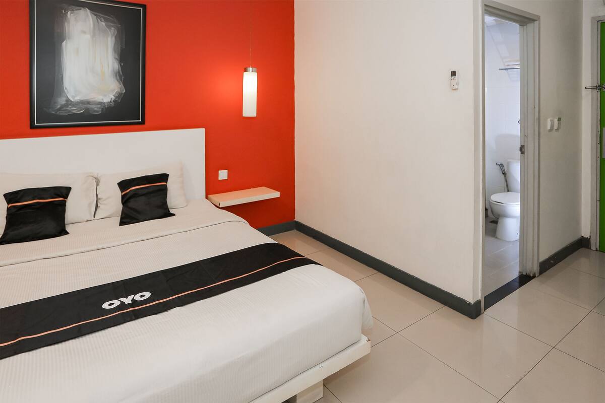 Habitación de hotel con cama, mesita de noche, luz de pared y puerta que da a un baño.