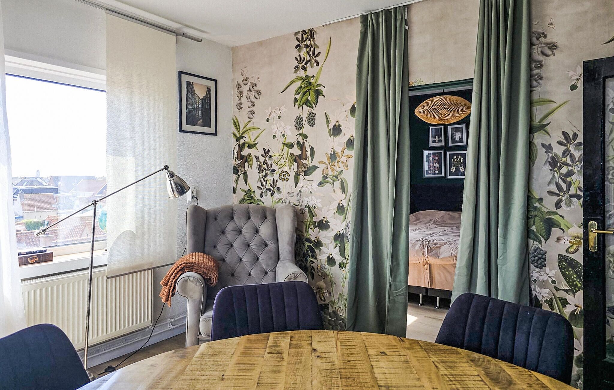 Una habitación con una butaca tapizada, una cama con cortinas verdes, una mesa de comedor y vistas a edificios a través de una ventana.