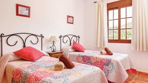 A room with two beds, each with a patterned bedspread and a red pillow. There is a nightstand with a lamp between the beds, and a window with curtains on the right side. - Casa Santiago (Frigiliana)
