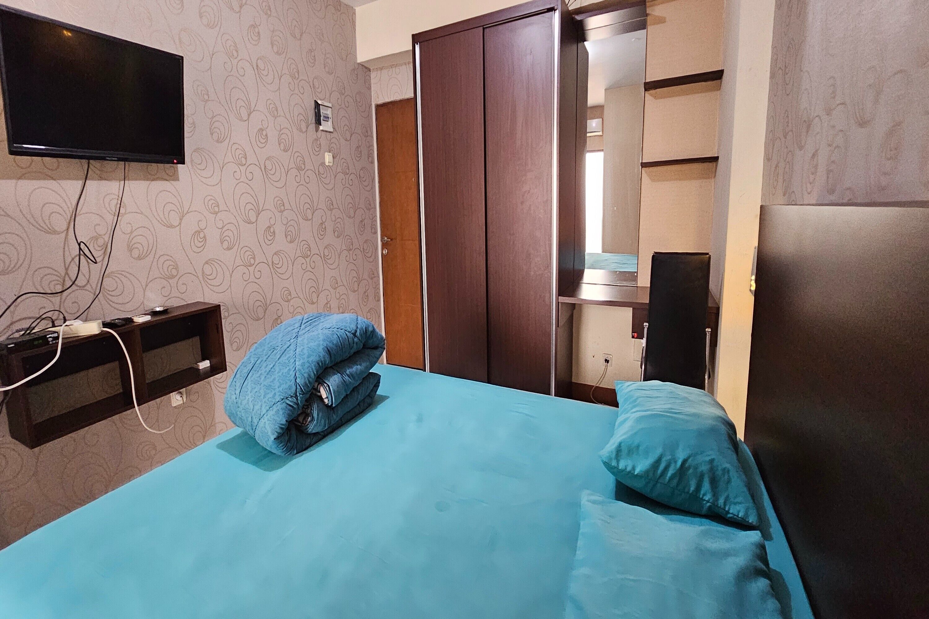 A hotel room with a bed, a TV mounted on the wall, a wardrobe, and a desk with a chair.