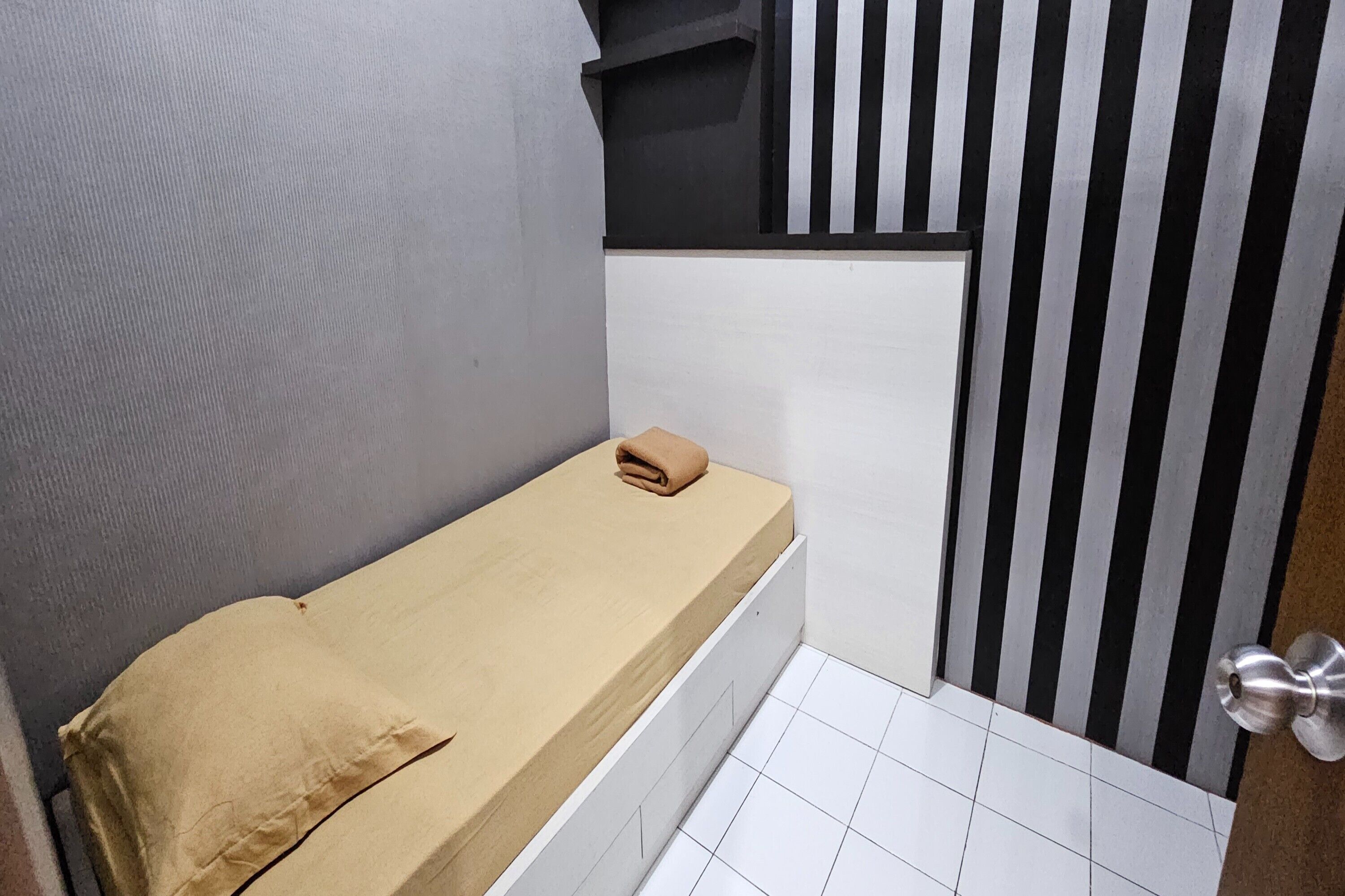 Quarto pequeno e estreito com uma cama de solteiro, uma toalha sobre a cama e uma porta com puxador metálico.