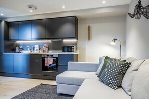 A modern kitchen with dark cabinets, a microwave, and a stove. There is a grey sofa with a polka dot pillow and a floor lamp.
