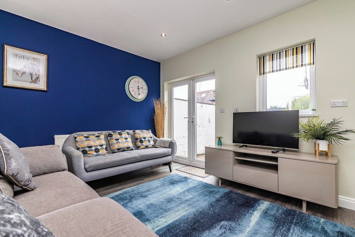 A living room with a sofa, a television on a stand, a clock, and a window with blinds.