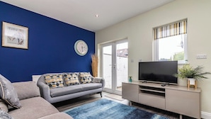 A living room with a sofa, a television on a stand, a clock, and a window with blinds.
