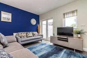 A living room with a sofa, a television on a stand, a clock, and a window with blinds.