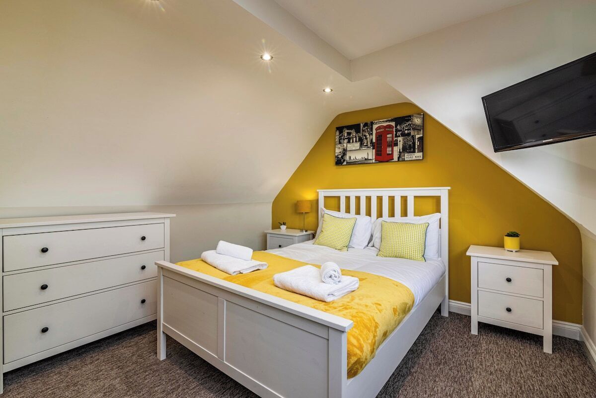 A bedroom with a bed, white nightstands, a flat-screen TV, and a yellow accent wall.