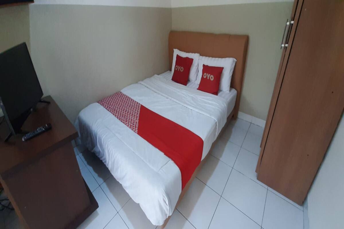 A hotel room with a bed, a wooden nightstand with a remote control, and a wardrobe.