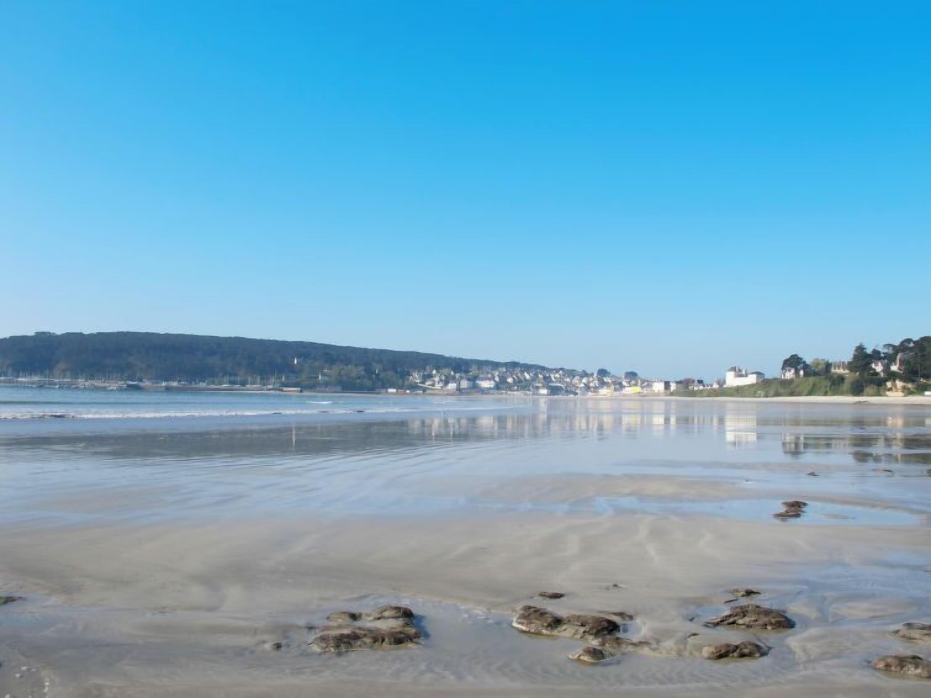 Une plage avec un ciel dégagé, une eau calme et une côte lointaine parsemée de bâtiments.