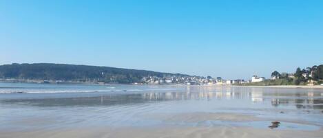 Une plage avec un ciel dégagé, une eau calme et une côte lointaine parsemée de bâtiments.