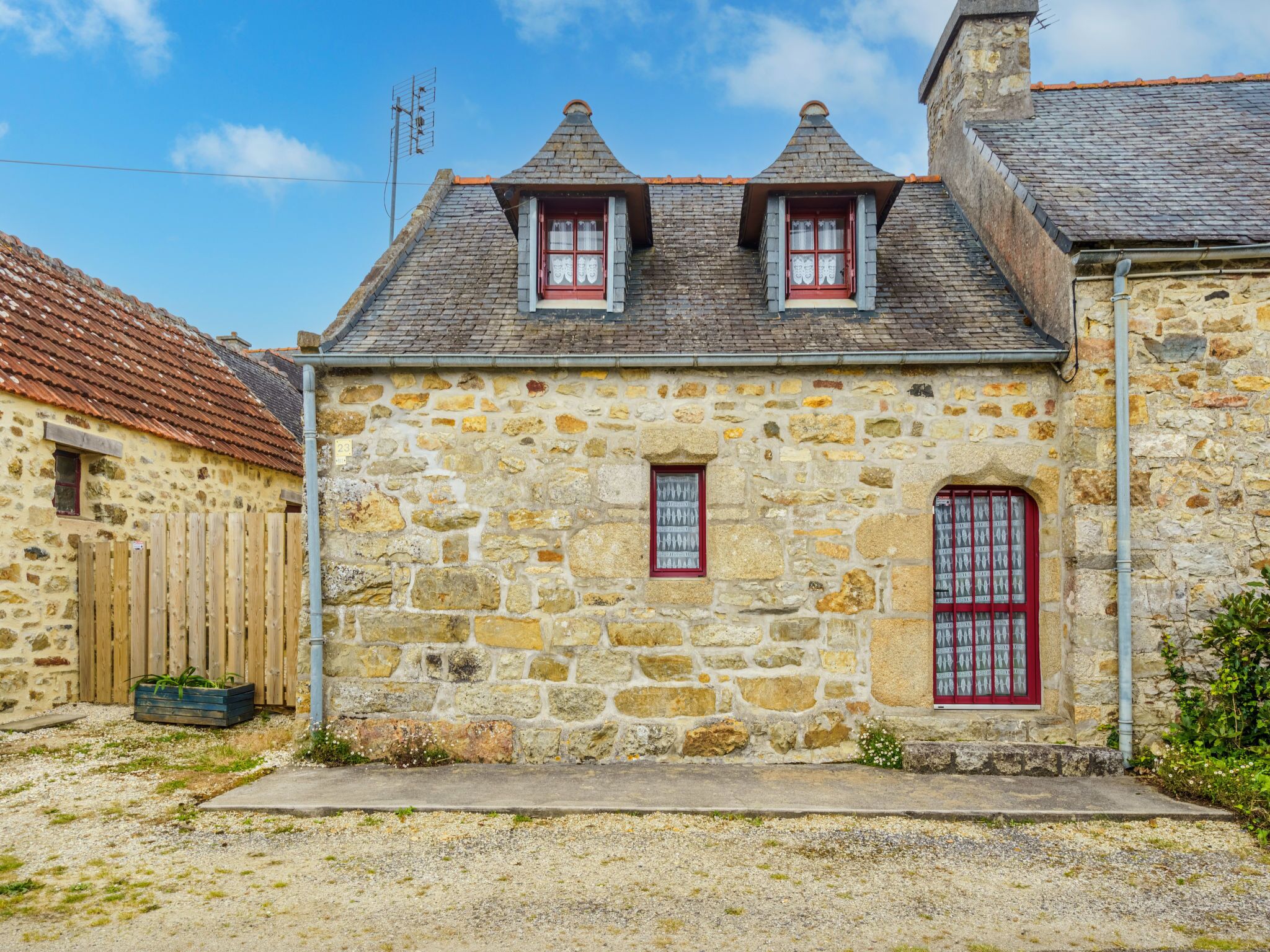 A stone building with two red-framed windows and a small door, surrounded by gravel and a wooden fence.
