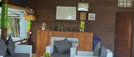 Amplia habitación con techo alto, paneles de madera y una mesa de recepción. Hay varios sofás de mimbre blanco y una silla de madera.