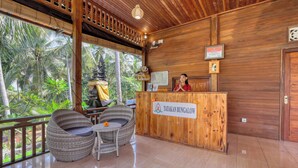 A reception area with a wooden reception desk labeled "TATAKAN BUNGALOW," wicker chairs, and a view of palm trees through large windows.
