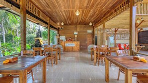 A wooden reception desk with a sign, surrounded by wooden tables and chairs in a spacious indoor area with a tiled floor and a view of greenery outside.