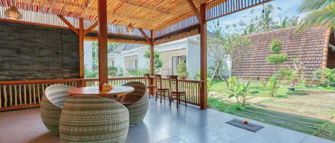 A covered outdoor seating area with wicker furniture and a view of a garden and buildings.