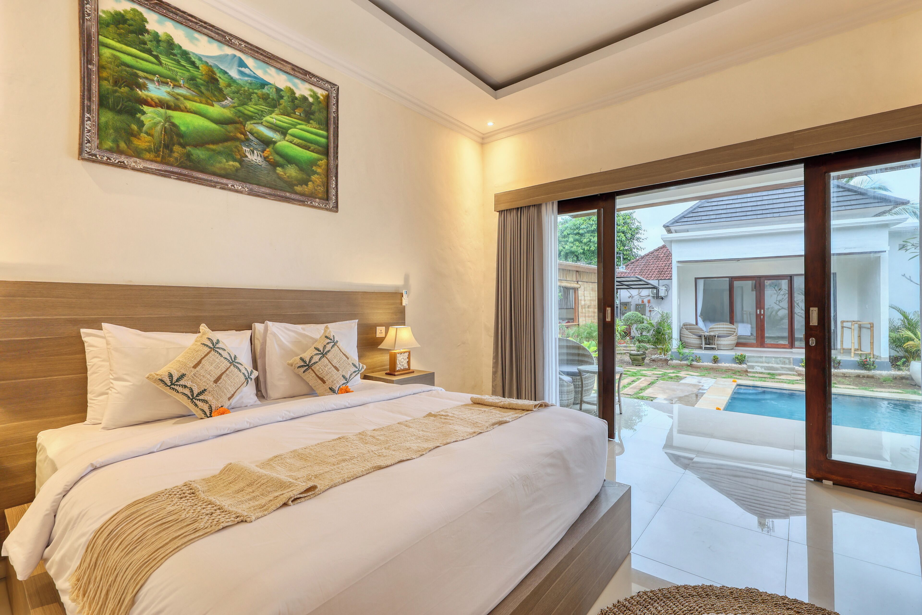 A hotel room with a large bed, a painting, a view of a swimming pool, and a balcony with outdoor furniture.