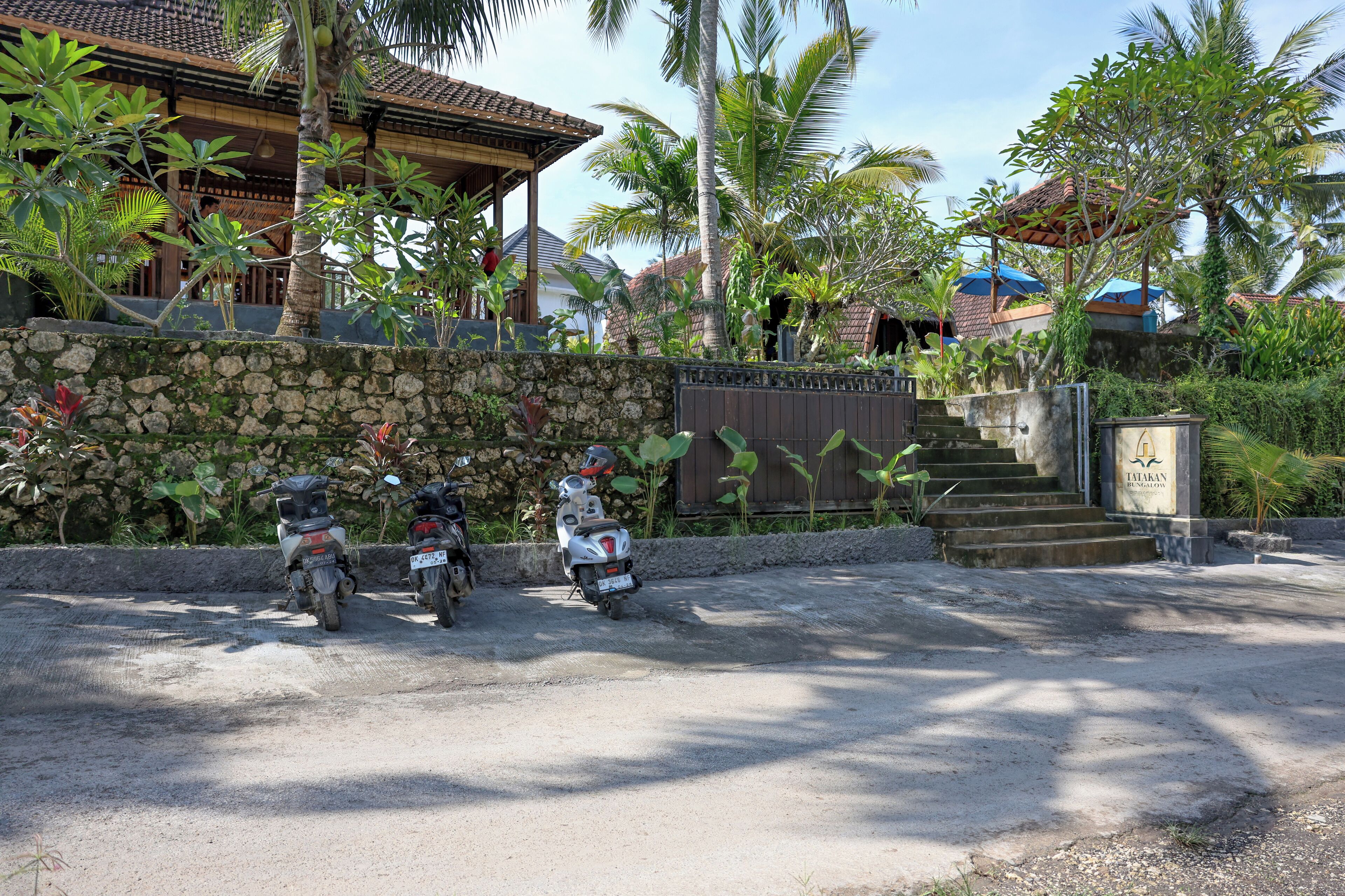 A stone wall with two parked scooters, a staircase leading to a building with a tiled roof, and lush tropical vegetation.
