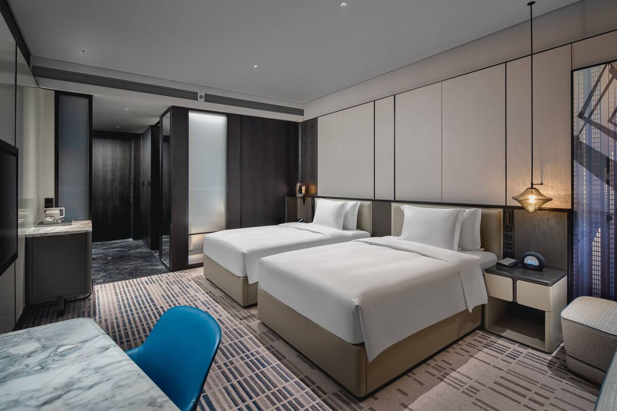 A hotel room with two beds, a desk, a chair, and a TV.