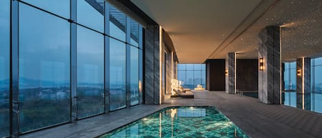 A modern indoor swimming pool with a tiled floor, large windows offering a city view, and a lounge area with seating.